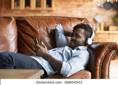 Calm young african ethnicity man in glasses wearing headphones, relaxing on comfortable couch listening favorite music, choosing tracks in online mobile application, casual hobby weekend lazy pastime.