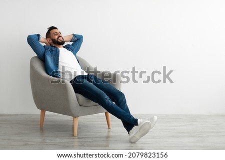 Calm young Arab man relaxing in armchair against white studio wall, copy space. Handsome middle Eastern guy enjoying peaceful weekend morning, resting with hands behind head