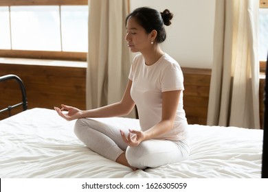 Calm young Asian woman sit on bad with mudra hands meditate with closed eyes in bedroom, relaxed millennial Vietnamese female practice yoga relieve negative emotions, stress free, meditation concept
