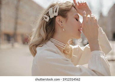 Calm young attractive blonde curly woman in pearl jewelry poses outside. Charming beautiful girl in white blouse smiles outdoors.