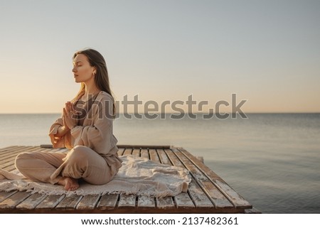 Calm young caucasian woman is meditating while sitting on wooden pier by sea. Brown-haired woman with her eyes closed tries to relax, breathes deeply and reaches nirvana.
