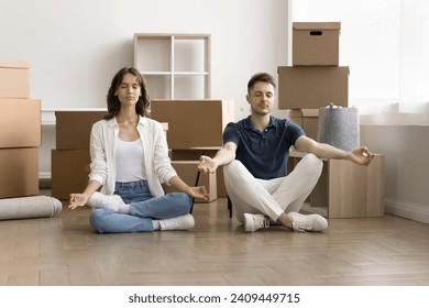 Calm young couple of new home owners meditating at paper moving boxes, sitting on floor, keeping zen hands and closed eyes, practicing stress relief after relocation activities