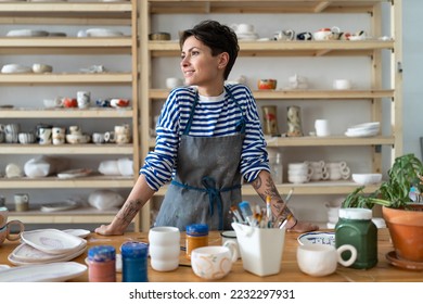 Calm young female owner in pottery workshop looking at window and smiling, plans projects. Small business concept, ceramics studio. Successful italian woman entrepreneur with tattoos at workplace.