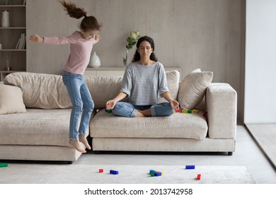 Calm Young Hispanic Mom Sit On Sofa At Home Meditate Practice Yoga Keep Calm Relieve Negative Emotions. Active Energetic Ethnic Daughter Jump Play Near Relaxed Mother. Motherhood Concept.