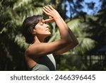 Calm young Hispanic woman holding hands in namaste meditating doing yoga breathing exercises with eyes closed feeling peace of mind, mental balance standing in green nature tropical park.