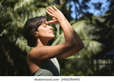 Calm young Hispanic woman holding hands in namaste meditating doing yoga breathing exercises with eyes closed feeling peace of mind, mental balance standing in green nature tropical park. - Powered by Shutterstock
