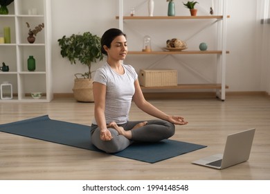 Calm young Indian woman sit on mat at home meditate training with online webcam class or lesson on computer. Millennial ethnic female practice yoga with distant web training session on laptop.
