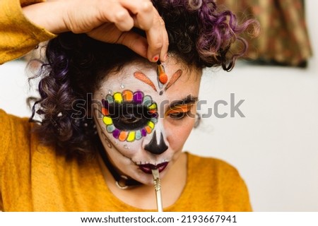 Calm young latin caucasian argentinian woman drinking mate in her room while applying La Calavera Catrina makeup with a small brush