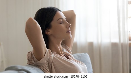 Calm young woman with closed eyes and hands behind head relaxing on couch close up, lazy day weekend at home, satisfied girl leaning back, stretching on sofa, daydreaming and meditating