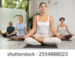 Calm young woman sitting in Lotus Pose Padmasana on mat during meditation and group yoga class in fitness studio