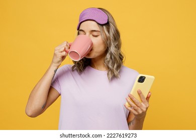 Calm young woman wears purple pyjamas jam sleep eye mask rest relax at home hold use mobile cell phone cup drink tea isolated on plain yellow background studio portrait. Good mood night nap concept