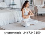A calm young woman in a white fitness suit sits in a lotus position and does yoga in the morning in the bedroom. A charming curly-haired girl enjoys stretching and working out at home. Copy space.