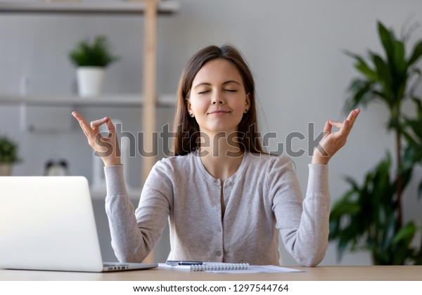 Calm young woman worker taking break doing yoga\
exercise at workplace, happy mindful female student meditating at\
home office desk feel balance harmony relaxation, stress relief zen\
at work concept