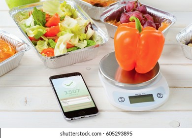 Calorie Counter App On The Smartphone, A Kitchen Scale And A Fresh Pepper On The Wooden Surface, Close-up. Lettuce And Tomato Salad, Grapes On The Background. Counting Calories.