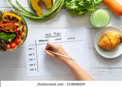 Calories control, meal plan, food diet and weight loss concept. top view of hand filling meal plan on weekly table with salad and fresh vegetable on dining table - Powered by Shutterstock