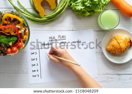 Calories control, meal plan, food diet and weight loss concept. top view of hand filling meal plan on weekly table with salad and fresh vegetable on dining table