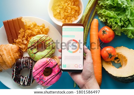 Calories counting and food control concept. woman using Calorie counter application on her smartphone with fresh vegetables, dessert and donuts on dining table
