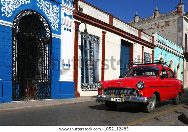 Camaguey, Cuba\
- March 7, 2018: Red classic car passes in front of a blue and\
white building. Same colors as cuban\
flag.