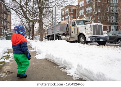 Cambridge, MA - January 28 2021: Young Kid Observing A Big Dump Truck Cleaning Up Snow In A US Neighborhood During Winter.