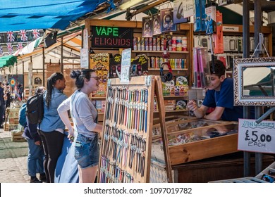 Cambridge, UK - Circa May 2018: Young Woman Seen Talking To A Shop Assistant Selling E-cigarette And Other Products In An Open Air Market In A City Centre. A Wide Variety Of Products Are Seen On Sale.