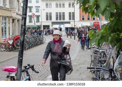 Cambridge, UK - Circa May 2019: Loan Adult Asian Woman Seen Walking Down A Pedestrian Only Area Within This Famous, Old City. Student Bikes And Other Members Of The Public Are Seen On A Busy Day.