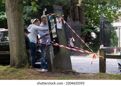 CAMBRIDGE, UK - SEPTEMBER 10 2022: Couple With Child Stands In Front Of A Crooked Parking Machine That Is Protected Against Falling Over By Red And White Ribbons. A Car Hit It