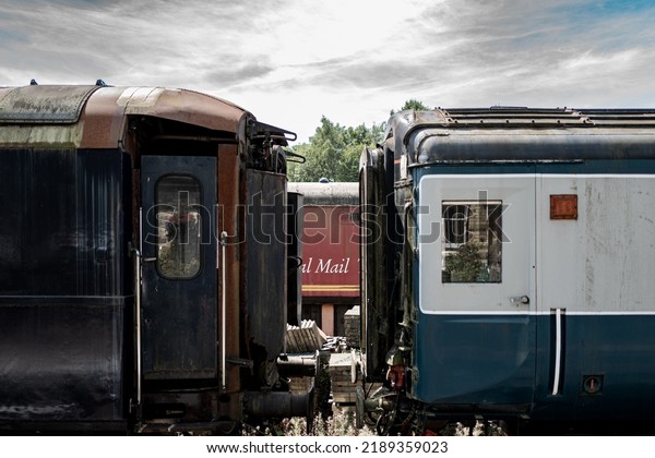 Cambridgeshire, UK -\
Circa August 2022: Shallow focus of a distant red Royal Mail train\
seen between old passenger carriages. Located in a shutting yard,\
they are not used\
anymore.