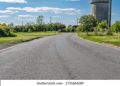 Cambridgeshire, UK - Circa May 2020: Ground level view of an empty rural road showing the textured road surface. A distant  Road Sign can be seen near a sharp bend, near a water tower.