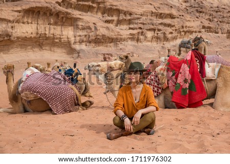 A camel driver in a cowboy hat and boots sits on the sand among the herd against a background of mountains. Shot in the Wadi Rum Desert in Jordan in March 2020.