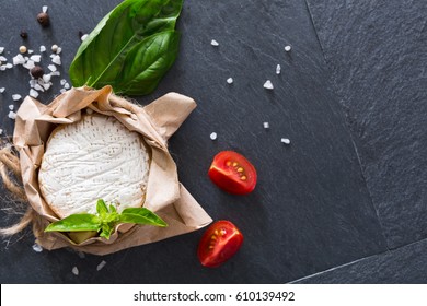 Camembert or brie circle cheese in brown kraft paper decorated with basil and pieces of cherry tomatoes, top view image with copy space on black stone desk surface
