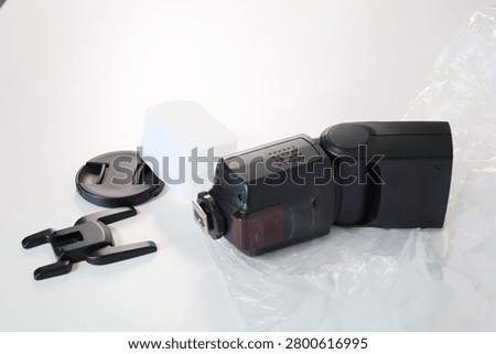 Camera flash, speedlight with diffuser, mini stand, and lens cap on a white background. A high-angle studio shot of a professional black external speedlite lying on a plastic wrap next to its included accessories, including a translucent white dome light modifier.