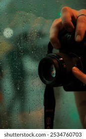 camera in focus next to a large window with raindrops and a person holding an umbrella silhouette.