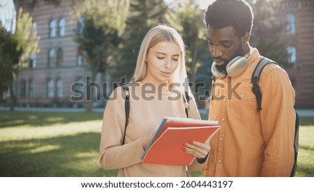 Camera moving closer to two students standing in front of big university. Girl showing something in her notebook to friend. Students spending break between lectures together. Sunny weather.