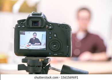 Camera On A Tripod Filming A Blogger At Home Sitting On His Workspace