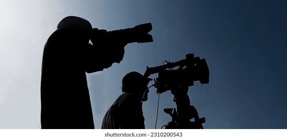 Camera operator on a tripod, photographer, cameraman silhouette isolated on blue sky background. 2 two men working together.  - Shutterstock ID 2310200543