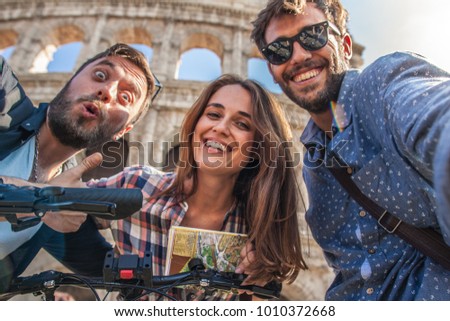 CAMERA POV Three happy young friends tourists with bikes and backpacks at Colosseum in Rome taking selfies pictures with smartphone having fun. Lens Flare.