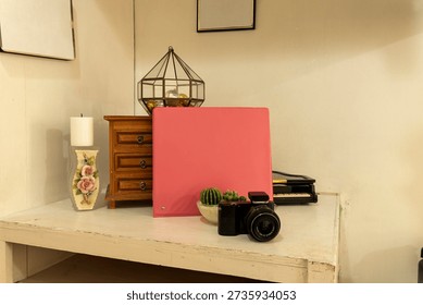 A camera sits on a table next to a pink folder and small plants. A wooden drawer and decorative items fill the background in a well-lit room.