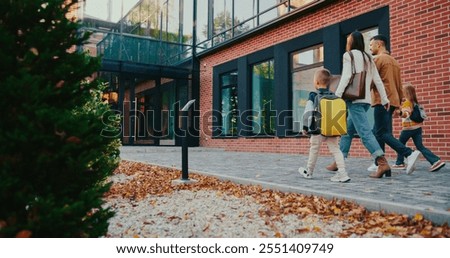 Camera view from behind small park area. Kids with backpacks running to school entrance. Parents waving their hands to children. Saying goodbye. Childhood concept. Yellow leaves on ground.
