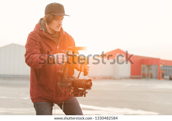 Cameraman with the camera\
and the stabilizer in his hands creates a video on the background\
of the sunset. Man takes a video with a stabilizer on the street.\
Backstage photo
