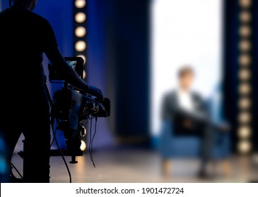 Cameraman filming in tv talk show studio. The host or presenter sitting on a chair camera pointed at him. Television news live broadcast production set