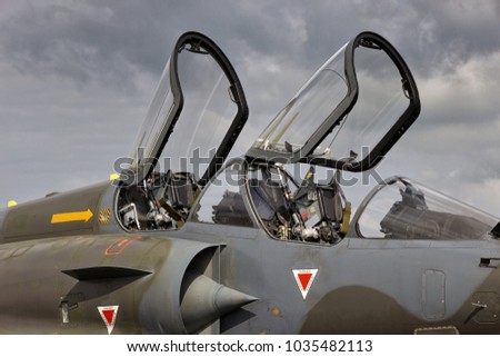 Camouflaged military fighter jet aircraft dual seat cockpit with open canopies.