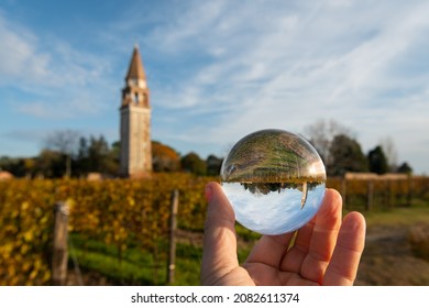 Campanile Di Sant Angelo On The Island Mazzorbo Near Burano (Venice, Italy) On A Sunny Day In Autumn, Colorful Leaves, Hand Holding Glass Ball