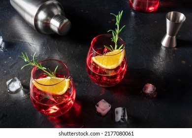 Campari Orange Cocktail With Mixing Tools, A Jigger And A Shaker, With Ice Cubes, And Rosemary For Garnish, On A Black Slate Background