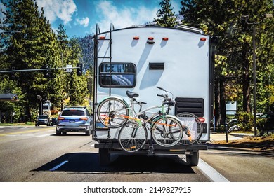 Camper With Two Bicycles Tied To Back On Tree-lined Highway Outside Of Lake Tahoe With Mountains In Distance On Sunny Summer Day