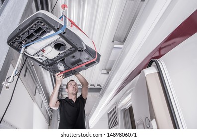 Camper Van Cooling Unit Installation By Professional RV Shop Technician. Caucasian Recreational Vehicles Service Worker Lifting Heavy Air Conditioner Unit On The Motorhome Roof.