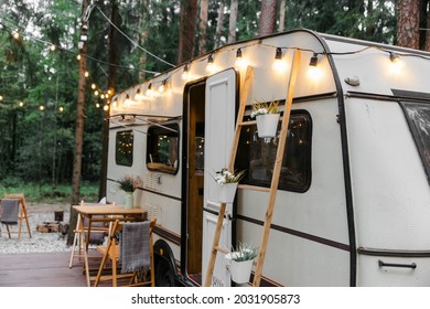 A camper van trailer with a wooden table and chairs. Decorative incandescent lamps on the street. Pine forest. Rest with the whole family in the country, fun trips in the fresh air. Picnic camping