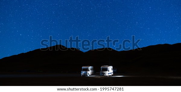 Campers parked in a caravan parking area sleeping on\
a starry night in the mountain. Summer tourism with RV in a blue\
night sky with stars. Best option for travel. Panorama motorhomes\
and camping car.