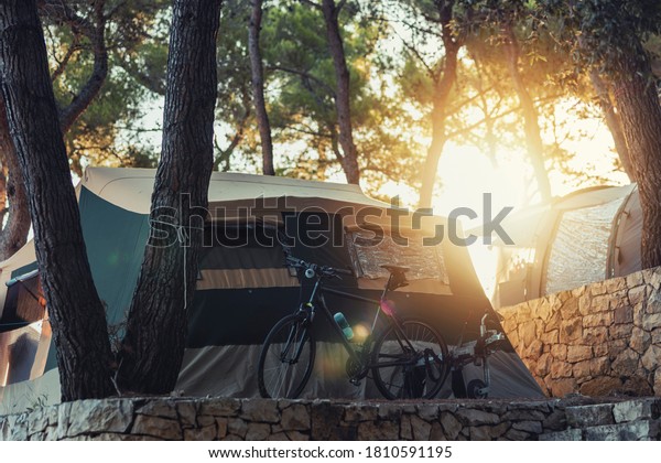 Campervan caravan vehicle home camper mobile motor\
home. Big comfortable tent resting in tree\'s shadow. A golden\
sunshine makes its way through tree′s leaves and sets light upon\
tent and bike.