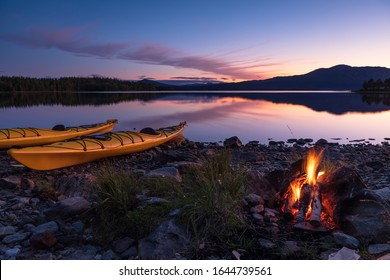 Campfire during dusk at the shore of a lake with two kayaks. Sweden.