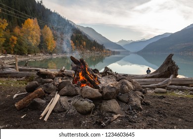 A campfire and kindling sit next to a rock and drift wood covered beach with the mountains and colourful fall leaves reflecting in the calm waters of Lillooet Lake in the background as the sun sets. - Shutterstock ID 1234603645